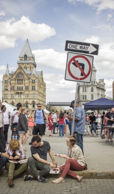 A line of for sit along a curb eating, while the festival masses can be see behind them.