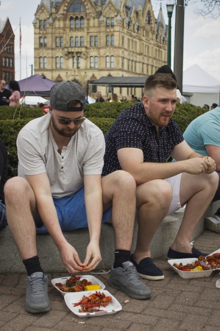 Two men sit along a curb on Clinton Square. Both have takeout containers of crawfish at their feet and are in the midst of eating them.