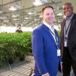 Two men stand off to the right smiling for a photo. Behind them are rows of marijuana plants being grown in a medical facility.