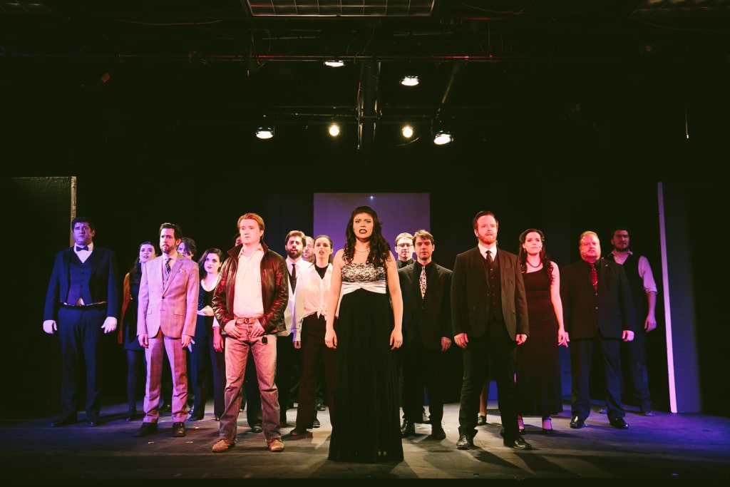 The cast of CNY Playhouse's "Chess" stand in a line across the stage as part of the performance, lit by overhead stage lights.
