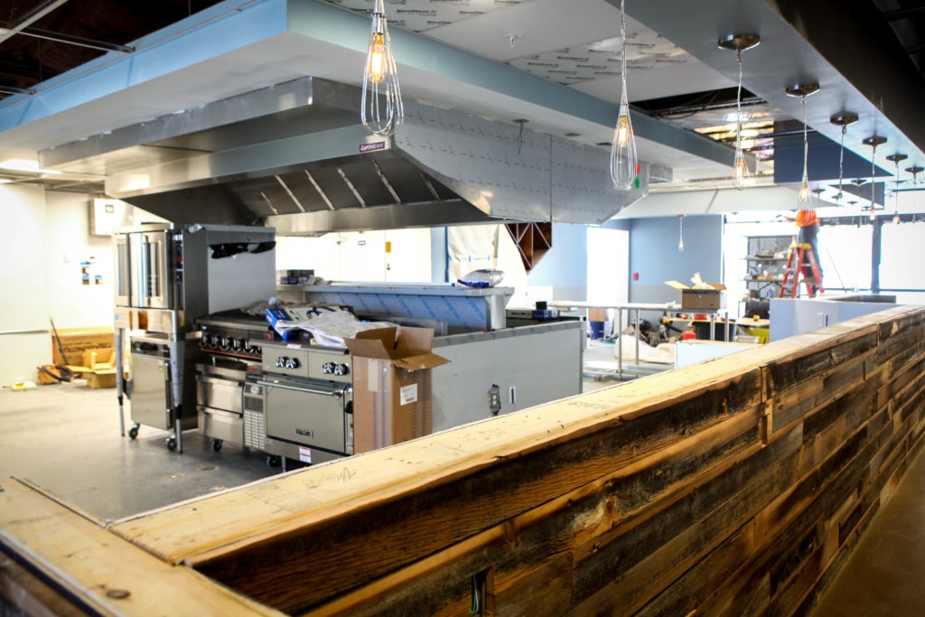 An under construction kitchen space at Defi Cuisine. The open steel stove is behind a wooden bar, with cardboard boxes scattered about.