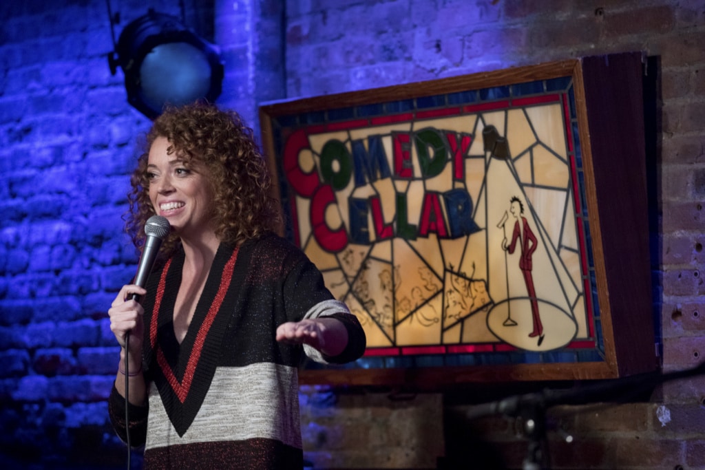 Comedian Michelle Wolf holds a microphone and does a comedy sketch on a stage.