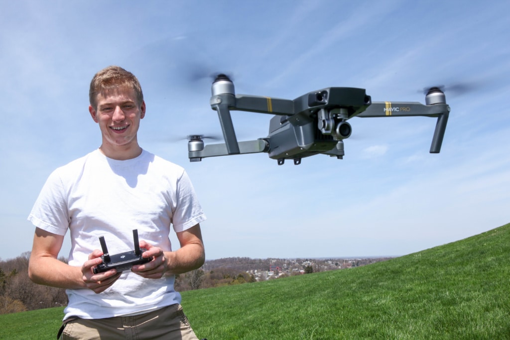 Chase Guttman stands to the left, holding a controler in his hands. A drone hovers in front of him while he controls it.