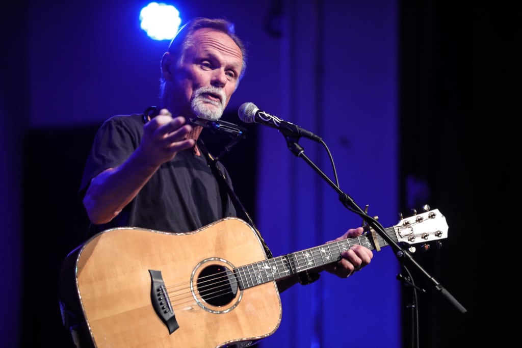 Jonathan Edwards stands on stage with an acoustic guitar, singing into a microphone.